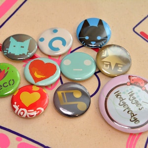 May include: A collection of ten circular pin-back buttons featuring various designs including a pixelated cat, a blue circle, a black cat face, a green flower with Neco Corporation text, a red heart, a light blue face, a gold star, a red heart, a gold musical note, and a light pink button with a pig illustration and the text Higgies Hodgepodge.