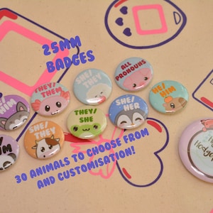 Puede incluir: Una colección de insignias de 25 mm con ilustraciones de animales adorables y varias opciones de pronombres, incluyendo "Él/Él", "Ella/Ella", "Ellos/Ellas" y "Todos los pronombres". Las insignias se muestran en una superficie marrón con un diseño de mando de juego rosa y azul. El texto "30 animales para elegir y personalizar!" también es visible.