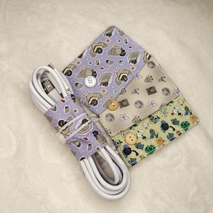 May include: A white USB cable wrapped with a lavender fabric cable organizer featuring a repeating pattern of cartoon animals and floral designs. Three additional fabric organizers with button closures are visible.
