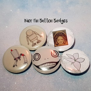 May include: Five Nier pin button badges. The badges are round and feature different characters from the game. One badge features a robot, another features a flower, another features a face with a large smile, another features a woman with long hair, and the last badge features a robot with a red light on its head.