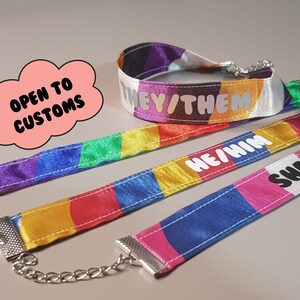 Könnte beinhalten: Drei bunte Satinband-Lanyards mit silbernen Ketten und Verschlüssen. Die Lanyards haben verschiedene Farben der Pride-Flagge und den Text "They/Them", "He/Him" und "She/Them" aufgedruckt. Der Text "Offen für Bestellungen" steht auf einer rosa Wolke.