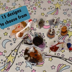 Op de afbeelding: Een verzameling van 15 verschillende karakterontwerpen op transparante acrylcharms en sleutelhangers. De charms tonen een verscheidenheid aan karakters, waaronder een kat, een hond, een vogel en een paddenstoel. De sleutelhangers zijn bevestigd aan zwarte koorden. De charms en sleutelhangers worden gepresenteerd op een witte achtergrond met een roze en blauw sterrenpatroon.