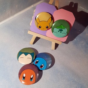 Peut inclure: Quatre boutons ronds représentant des personnages de dessins animés de la franchise Pokémon. Les boutons sont vert, bleu, orange et jaune. Les personnages sont Bulbizarre, Carapuce, Salamèche et Pikachu.