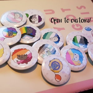 Op de afbeelding: Een verzameling van 13 ronde knoppen met een cartoon schaap met verschillende gekleurde wol die verschillende pride-vlaggen voorstellen. De tekst "Open to customs!" is zichtbaar op de achtergrond.