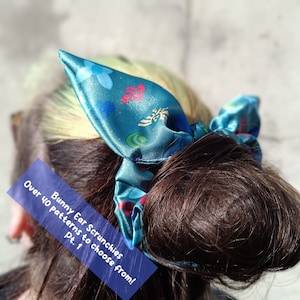 Op de afbeelding: Een tealblauwe satijnen scrunchie met een bloemen- en geometrisch patroon. De scrunchie is gebonden in een strik en wordt gedragen in het haar van een persoon. De tekst "Over 40 patterns to choose from! Pt. 1" is zichtbaar in de afbeelding.