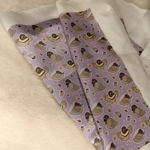 May include: A close-up of fabric with a repeating pattern of cartoon pigeons in various poses on a light purple background. The pigeons are gray, brown, and cream-colored. The fabric is folded over a white surface.