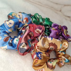 May include: A collection of five satin hair scrunchies in blue, red, green, purple, and gold. Each scrunchie features a repeating pattern of stylised cartoon character faces with various hair colours and expressions printed on the smooth fabric.