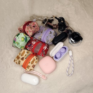 May include: An assortment of small, patterned fabric pouches with zippers, various earbuds, and charging cases. The pouches feature designs like floral, animal, and abstract patterns. Earbuds and cases are in white, black, and pink.
