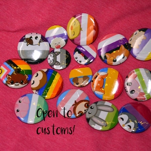 Op de afbeelding: Een verzameling van 16 ronde knoopsluitingen met verschillende dierillustraties met regenboogvlagontwerpen. De tekst "Open to customs!" staat onder de knoopsluitingen.
