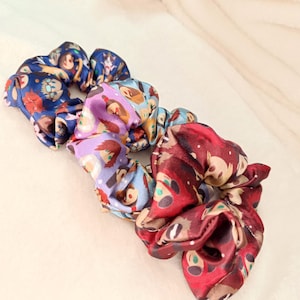 Könnte beinhalten: Drei bunte Haargummis mit einem Cartoon-Charakter-Druck. Die Haargummis sind blau, lila und rot.