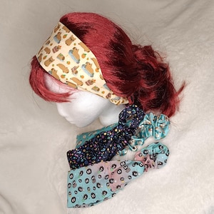May include: A cream-coloured headband with a capybara and orange print worn on a mannequin head, accompanied by three fabric scrunchies featuring patterns of colourful question marks, small cartoon soot sprites, and various character illustrations.