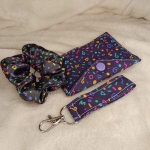May include: A black satin scrunchie, a small pouch, and a keychain wristlet. All items feature a dark purple background with colorful confetti-like shapes. The pouch has a purple button closure, and the keychain has a silver clasp.