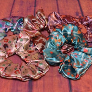 May include: Five colourful satin scrunchies with different patterns. The scrunchies are in shades of pink, blue, brown, and purple. Each scrunchie has a different pattern, including flowers, leaves, and animals.
