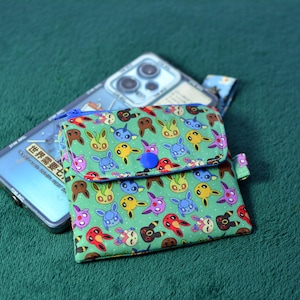 Pode incluir: Uma carteira de tecido verde com aba, adornada com um padrão colorido de personagens de desenhos animados. A carteira é presa com um botão azul e repousa sobre uma capa de telefone. A capa do telefone tem texto.