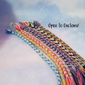 Pode incluir: Uma coleção de pulseiras de amizade coloridas com uma variedade de padrões. As pulseiras são feitas com fio de bordar e são amarradas em um padrão chevron. O texto "Open to Customs!" é visível na imagem.