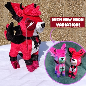 Op de afbeelding: Een rood, zwart en wit pluche speelgoed met een kruisontwerp en een bliksemschicht op het oog. De afbeelding bevat een kleinere inzet die een neonroze variant van het speelgoed laat zien. Tekst luidt "WITH NEW NEON VARIATION!"