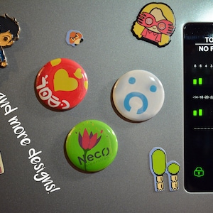 May include: A collection of assorted decorative magnets featuring various characters, symbols, and logos, including a red button with a yellow heart, a white button with a blue icon, and a green button with a flower logo, alongside the text and more designs.