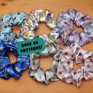 May include: Six colourful hair scrunchies with different patterns. The scrunchies are made of satin and have a variety of designs, including eyes, flowers, and cartoon characters. The text "Over 40 Patterns!" is written on a green tag.