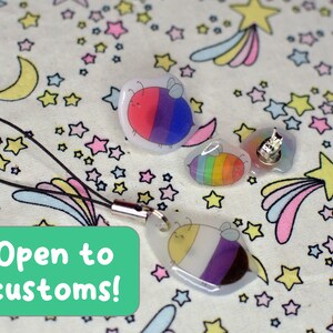 May include: Three resin bee charms with different pride flag designs. The charms are on a white background with a pattern of stars and shooting stars. The text "Open to customs!" is on a green rectangle in the lower left corner of the image.