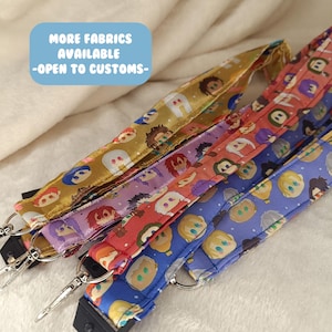 May include: A collection of colorful lanyards with various character designs. The lanyards feature a variety of colors, including gold, purple, red, and blue. Each lanyard has a metal clasp and a black plastic piece. The text "MORE FABRICS AVAILABLE -OPEN TO CUSTOMS-" is in a blue box.