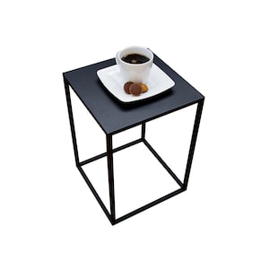 May include: A small, black square side table with a minimalist design. A white square plate with a cup of coffee and two cookies sits on top. The table has a simple, open frame.