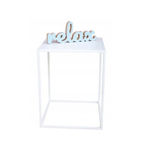 May include: White metal side table with a square top and a single shelf. The word "relax" is written in blue on a wooden sign on top of the table.