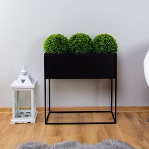 Puede incluir: Un macetero de metal negro con tres plantas artificiales redondas de color verde. El macetero est&aacute; sobre un soporte de metal negro con un estante inferior. El macetero est&aacute; delante de una pared blanca con una impresi&oacute;n enmarcada de un flamenco rosa.