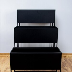 Puede incluir: Un soporte para plantas de metal negro con tres niveles. Cada nivel es una jardinera rectangular. El soporte est&aacute; sobre un suelo de madera.