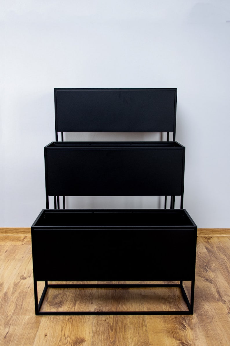 Puede incluir: Un soporte para plantas de metal negro con tres niveles. Cada nivel es una jardinera rectangular. El soporte est&aacute; sobre un suelo de madera.