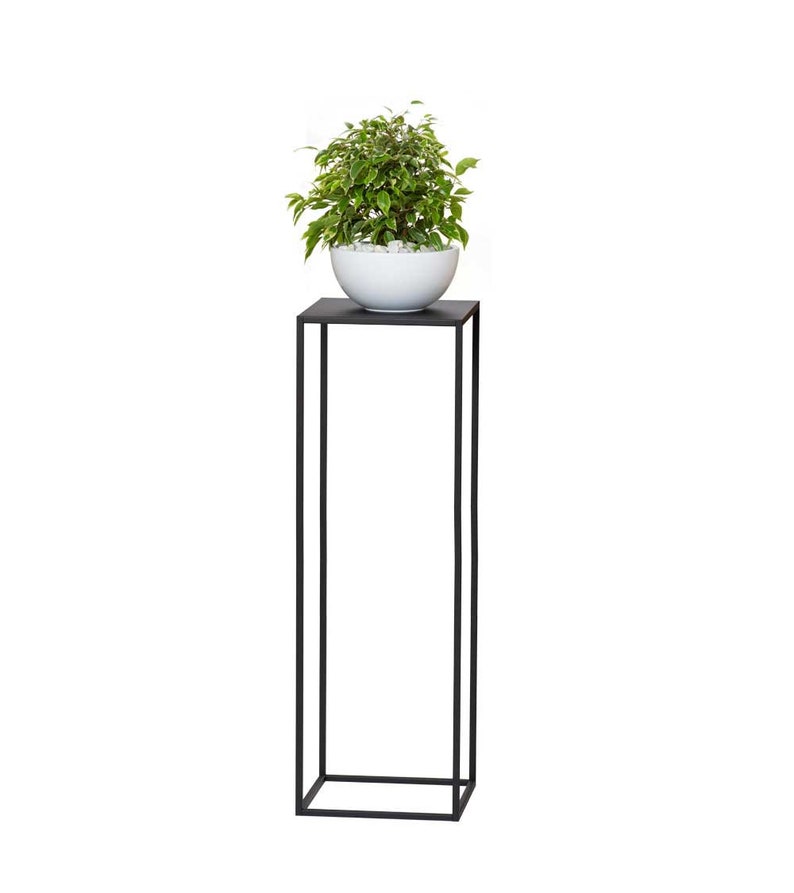 May include: A modern black metal plant stand with a square top and a tall, rectangular base. A white ceramic bowl filled with small white stones and a green and white leafy plant sits on top. The stand is a stylish way to display plants.