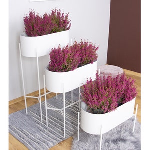 May include: Three white oval planters with pink flowers are stacked on a white metal stand. The stand has three tiers and is on a grey and white striped rug. A grey fluffy rug is in the foreground.
