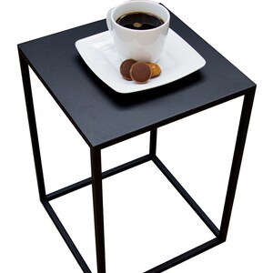 May include: A black square side table with a minimalist design. A white square plate holds a white coffee cup filled with dark liquid and two chocolate candies. The table has a geometric, open frame.