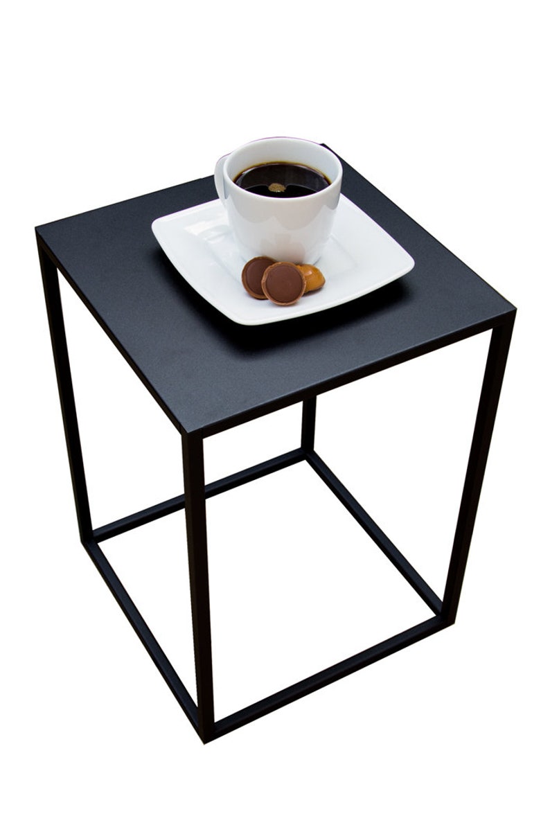 May include: A black square side table with a minimalist design. A white square plate holds a white coffee cup filled with dark liquid and two chocolate candies. The table has a geometric, open frame.