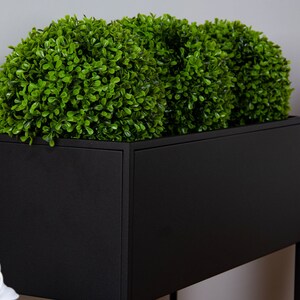 Puede incluir: Un macetero de metal negro con tres bolas de topiario artificiales verdes. El macetero est&aacute; sobre un soporte de metal negro.