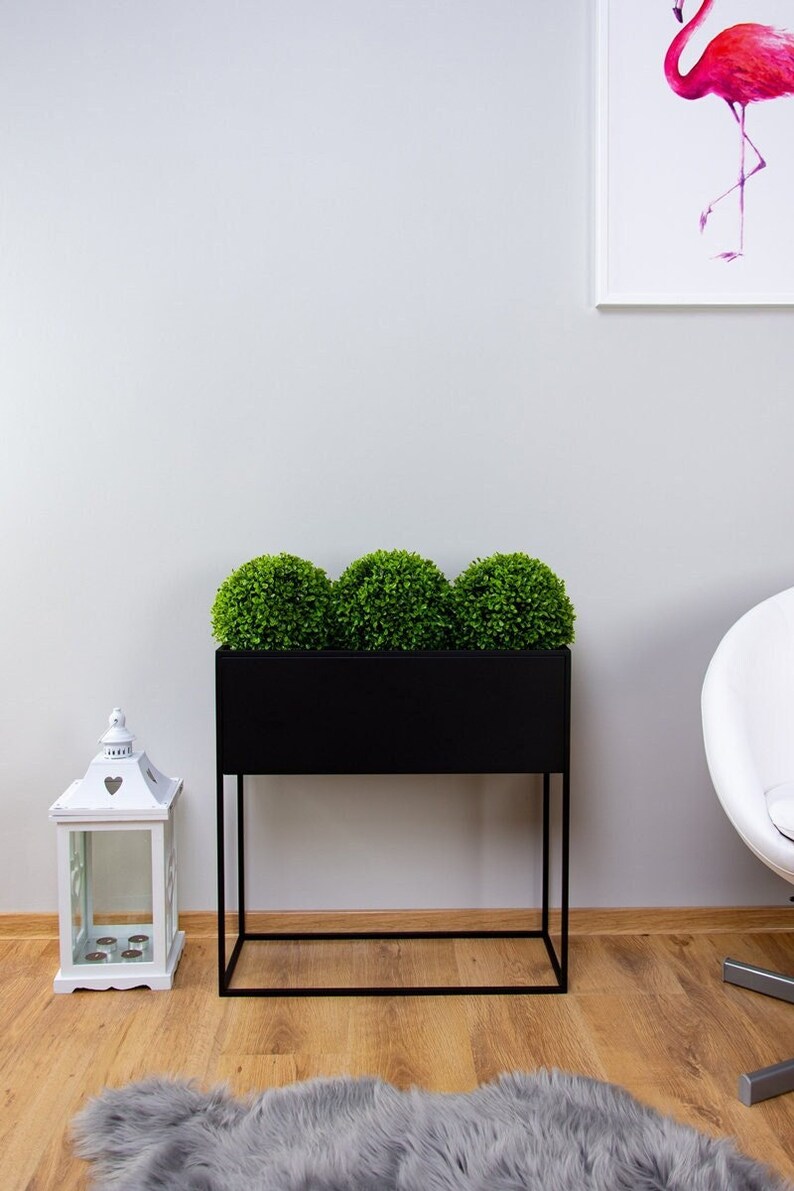 Puede incluir: Un macetero de metal negro con tres plantas artificiales redondas de color verde. El macetero est&aacute; sobre un soporte de metal negro con un estante inferior. El macetero est&aacute; delante de una pared blanca con una impresi&oacute;n enmarcada de un flamenco rosa.