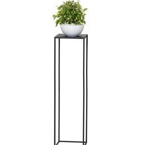 May include: A tall, black metal plant stand with a square top supporting a white ceramic bowl filled with small white stones and a green and white leafy plant. The stand has a minimalist, modern design.