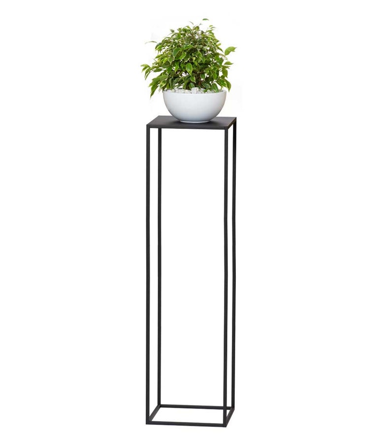May include: A tall, black metal plant stand with a square top supporting a white ceramic bowl filled with small white stones and a green and white leafy plant. The stand has a minimalist, modern design.