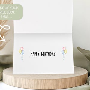 May include: A white birthday card with the words "HAPPY BIRTHDAY" in black. Watercolor balloons in pastel colors are on each side of the text. The card is displayed on a wooden stand.