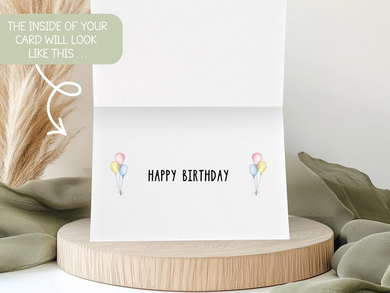 May include: A white birthday card with the words "HAPPY BIRTHDAY" in black. Watercolor balloons in pastel colors are on each side of the text. The card is displayed on a wooden stand.
