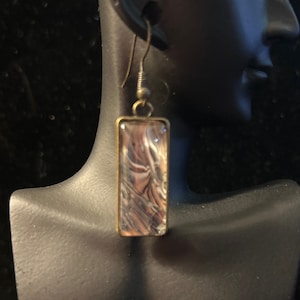 May include: A pair of gold-toned earrings with a rectangular, clear glass pendant featuring a swirling white and gray abstract design.