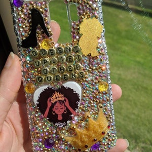 May include: A clear phone case embellished with iridescent rhinestones, gold, and purple accents. The case features a black high heel, a gold silhouette, and the text "QUEENS UPLIFT OTHER QUEENS". A graphic of a woman wearing a crown is also visible.