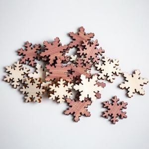 May include: A collection of wooden snowflake ornaments in natural and reddish-brown tones. The snowflakes are intricately cut with six points, perfect for holiday crafts or decorations. The ornaments are scattered on a white surface.
