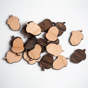May include: A collection of wooden acorn shapes in brown and natural wood tones. The acorns are scattered on a white surface.