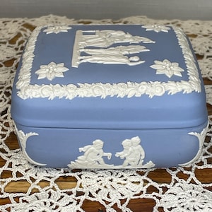 May include: A square, light blue trinket box with white relief detailing. The lid features a floral border and a central scene with figures. The box's sides have a curved design with more white relief figures. A decorative item.