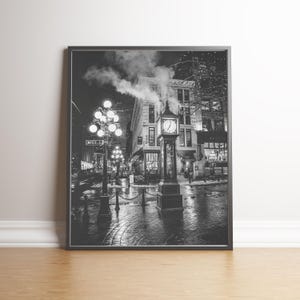 May include: Black and white framed print of a city street at night. A tall clock tower stands in the center, with a building behind it. Streetlights illuminate the wet cobblestone street. Steam rises from a building.
