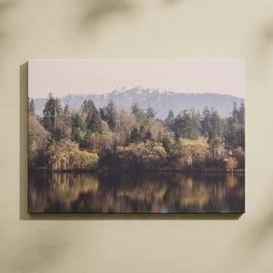 May include: A canvas print depicting a tranquil lake scene with mountains in the background. The image shows a forest with a mix of trees, including evergreens and deciduous trees with golden leaves. The water reflects the trees and sky, creating a peaceful atmosphere.