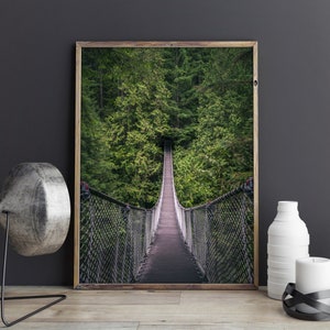 May include: A framed photograph of a wooden suspension bridge extending into a lush green forest. The bridge is made of wood and has metal mesh railings. The forest is dense with tall evergreen trees. The photograph is in a wooden frame.