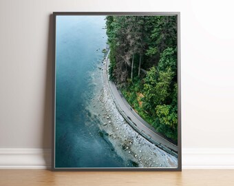 May include: An aerial view of a winding road along a rocky coastline with a lush green forest on the right side of the image. The water is a deep blue color and the sky is overcast.