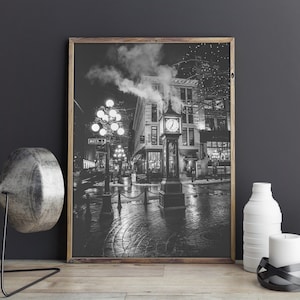 May include: Black and white photograph of a clock tower in a city street at night. The clock tower is illuminated by streetlights and has a large clock face with Roman numerals. The street is wet from rain and there are buildings on either side of the street.