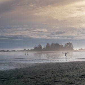 Puede incluir: Una figura solitaria camina por una playa de arena llevando una tabla de surf, con una peque&ntilde;a isla en la distancia y un cielo nublado arriba.